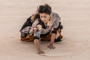 Foto do espetáculo "Ariadne: Cartografias de um Labirinto", mostra uma atriz, vestindo roupas escuras e com o cabelo assanhado, ajoelhada em um cenário desértico, arrastando suas mãos na areia.